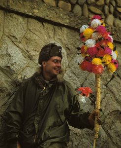 Korean War-Era American Soldier wearing an olive-drab military winter coat with brownish fur-lined hood around the collar. He is wearing a cold weather military flyers hat with synthetic fur lining. In his left hand he is holding a bouquet of pastel-colored paper flowers.
