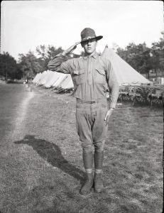 Balck and white photo of a World War I-Era Wisconsin National Guard soldier standing at the position of attention in front of a row of tents.