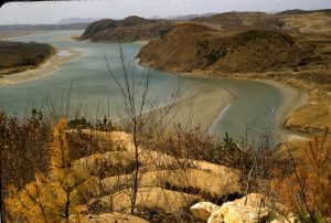 A color photograph of a Korean landscape featuring a winding body of water enclosed by greenish-brown hilly banks. There are large rocks in the foreground with coniferous trees popping up.
