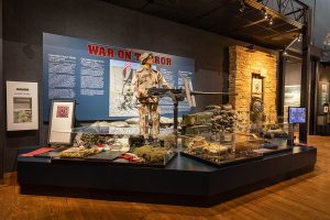 Woman at a machine gun dressed in desert fatigues overlooks artifacts from the OIF/OEF.