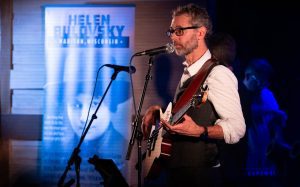Image of man with glasses and beard playing a guitar and singing on stage.