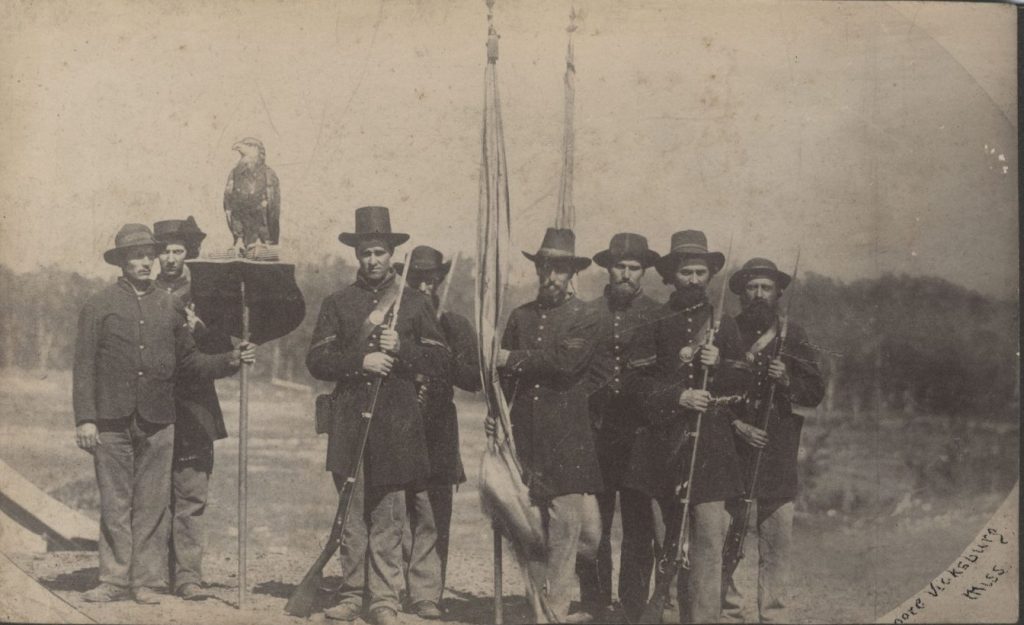 8 Civil War soldiers with two flags and an eagle on a stand posing for a photograph.
