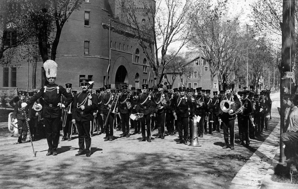 A group of people in uniform, carrying musical instruments on the street in 1910.