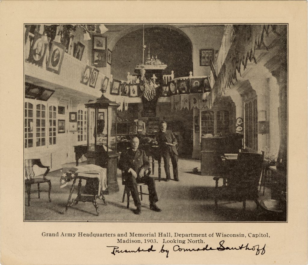 One men standing, one seated in a library-style room with portraits and artifacts..