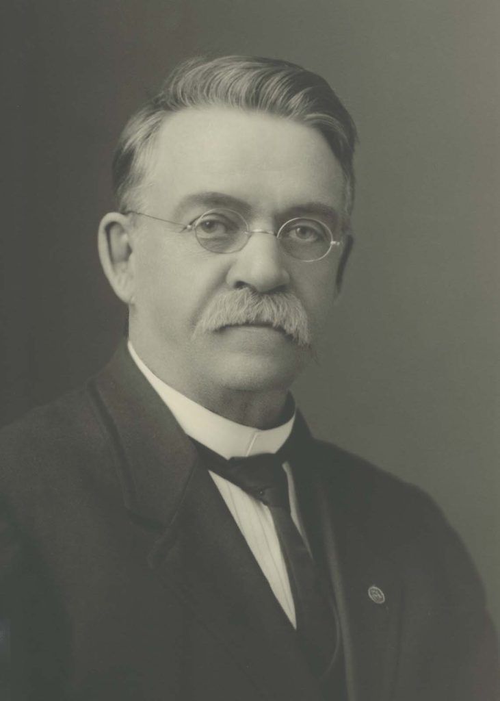 early 20th c. bust image of a middle-aged man with a moustache and glasses in a suit.