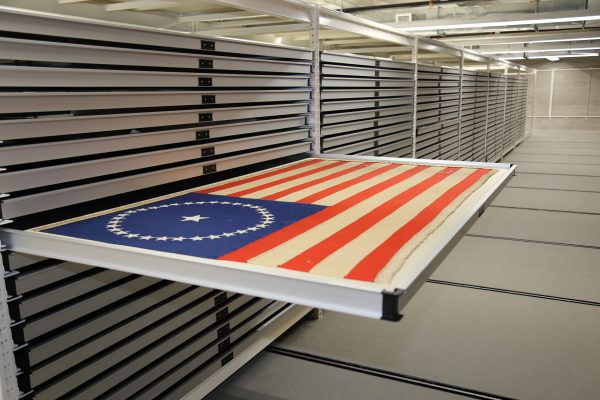 shelving rows with one open to show a US Civil War battle flag with red and white stripes and white stars on a blue field.