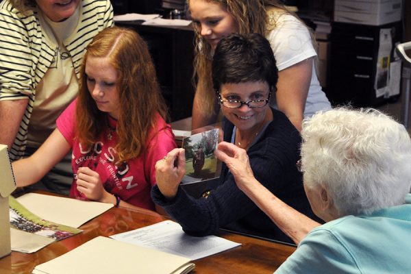 Three adult women and two younger women are gathered around a table with photographs and paperwork.