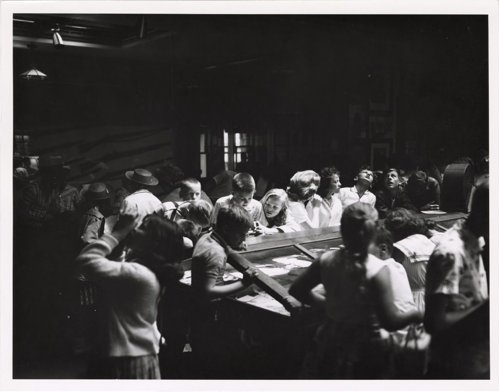 black and white photo of children gathered around cases in a museum.