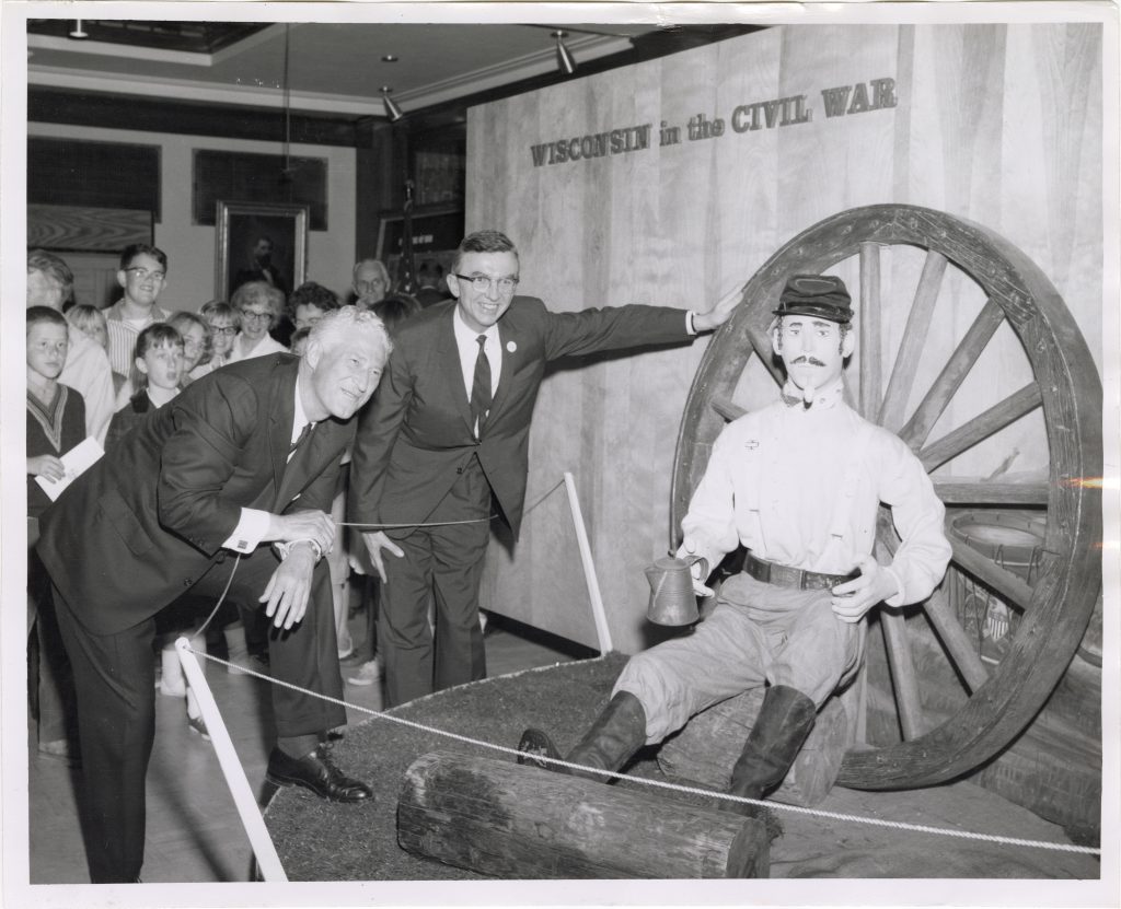 a mannequin sits in front of a wagon wheel in an exhibit with two men in suits smiling.