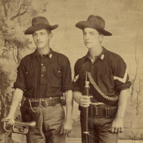 Two soldiers stand in front of a photography studio backdrop painted with foliage, wearing campaign hats with brass insignia, sack coats, uniform trousers with belts, and leather boots. 