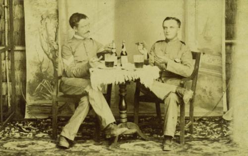 Two soldiers seated at a table, smoking cigars, playing cards and drinking. 