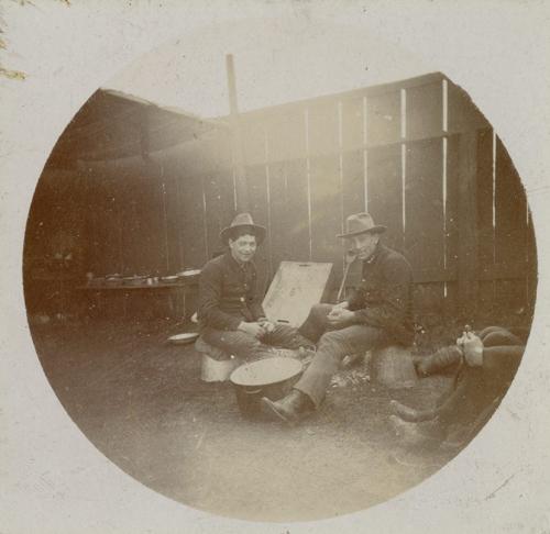2 soldiers sit on overturned buckets peeling potatoes over a pot. Both wear campaign hats, sack coats, uniform trousers, and boots.
