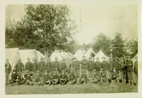 unidentified group of soldiers facing the camera in two rows, with the front row sitting and the back row standing. The soldiers wear campaign hats, uniform trousers, and boots. 