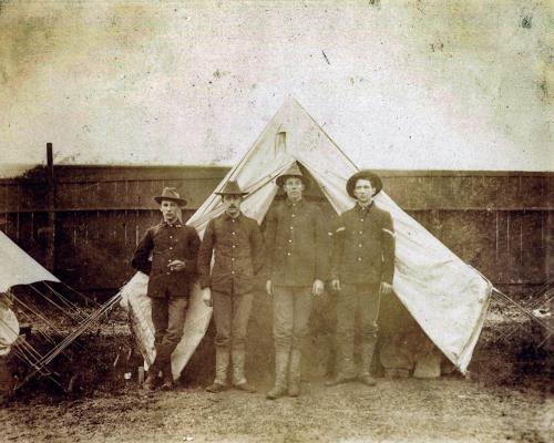 4 soldiers in front of a tent wearing campaign hats, sack coats, uniform trousers, and boots. 